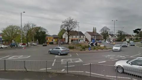 Traffic can be seen at the island on the main route in the city. There are buildings in the background along the road and some trees.