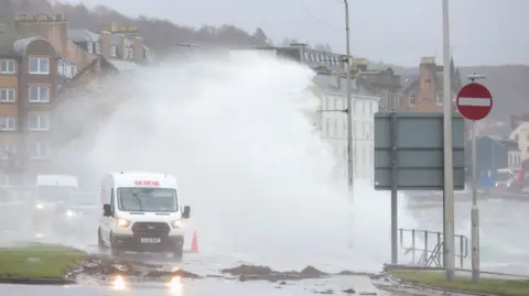 Reuters Two vans and a car drive through water crashing over the harbour wall in Oban