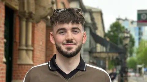 A young man with short brown hair, dark moustache and beard. He is wearing a green polo shirt. He has sunglasses on his head.