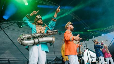 Four men wearing colourful military-style uniforms on a stage, with a variety of brass instruments.