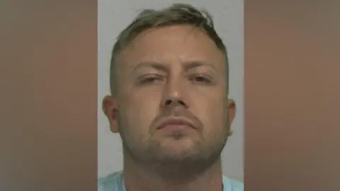 Mugshot of Watson. He has faint stubble and short blond hair.