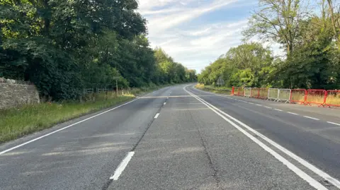 The old road to and from Birdlip in Gloucestershire. It is deserted with no traffic as it is now closed. There are trees either side of the dual carriageway and red and white metal fences blocking it off on the side. 