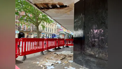 Owen Sennitt/BBC Debris can be seen on the floor on the pavement with red security fencing erected after parts of the canopy of the Debenhams department store in Norwich fell off.