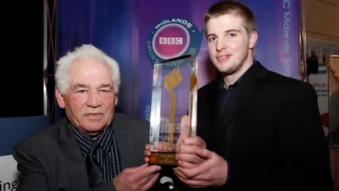 Two people standing indoors at an event booth display branded ‘BBC Midlands’ in the background. Both individuals are holding a tall, transparent trophy featuring a golden microphone design mounted on a base with engraved text. The backdrop includes a purple gradient panel with a circular BBC Sports logo and vertical ‘BBC Midlands’ text along the side. The setting appears to be an awards or media event, with exhibition stands and signage visible in the background.