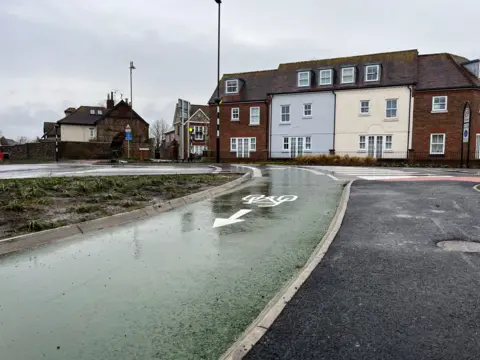 George Carden/BBC The green cycle lane which goes around the roundabout's perimeter. It's reflecting the sky as it has been raining. There is an icon of a bike and arrow painted on the lane in white. In the background are houses and one of the crossing points