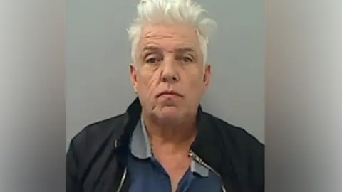 NCA Peter Lamb with white hair that is sticking up looks straight at the camera. He is wearing a blue open t-shirt with a black zipped jacket.