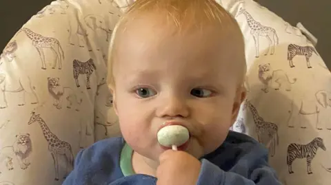 Archie Squire is sucking a lollipop sitting in a high chair wearing a blue top