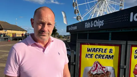 A man in a pink polo shirt looks at the camera. He is standing next to signs advertising waffle and ice cream. A big wheel is in the distance. 