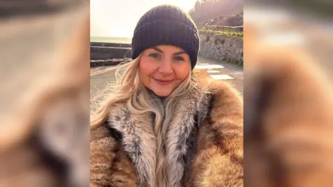 A midshot of Gemma in the winter sunshine, smiling to camera and wearing a big fluffy winter coat and beanie hat with a field and woods behind her