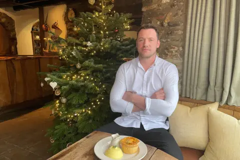 Olivia Richwald/BBC Tommy Banks, pictured in front of a Christmas tree with a plate of pie and mash in front of him. Mr Banks is wearing a white shirt and dark trousers and is sitting with his arms crossed.