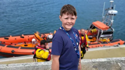 A young boy with brown hair wearing a blue shirt with the RNLI logo on it. In the background are two men wearing life jackets and an orange lifeboat on the water. 