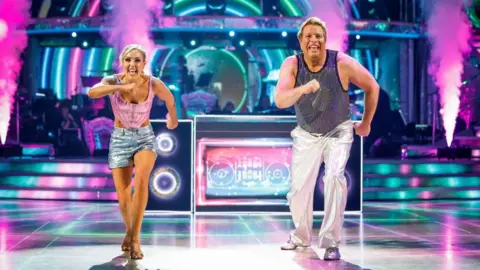 BBC/PA Amy Dowden is dancing alongside Thomas Skinner on the Strictly dancefloor. She is wearing a pink sleeveless top and silver hot pants and Skinner is wearing silver trousers and a sparkly silver top. A prop of a mock set of DJ decks is behind them