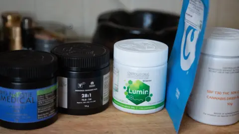 White and black plastic medical containers lined up on a wooden counter top. Some have blue and green coloured labels stating the containers carry medical cannabis. 