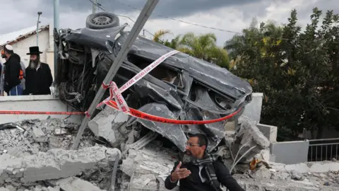 Man sitting beneath wrecked car gestures with his hand