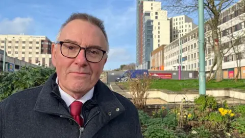 Plymouth City Council leader Tudor Evans in Armada Way in Plymouth. He is wearing a white shirt, red tie, grey coat and black-rimmed glasses. 