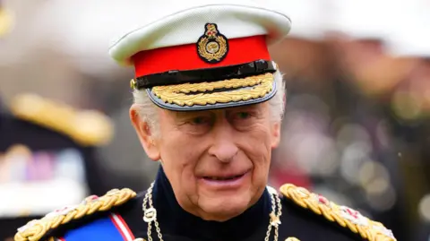 PA Media King Charles III in formal military uniform speaks to Royal Marines Commando personnel while they are lined up during a ceremony at Windsor Castle. He is wearing a white and red military hat.