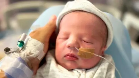 Lauren Farrelly Nico is a newborn baby lying on a hospital support surface, wearing a hat and a zip-up outfit, with a medical tube secured to the face and a monitoring line attached to the hand. Lauren's hand supports the baby’s head.