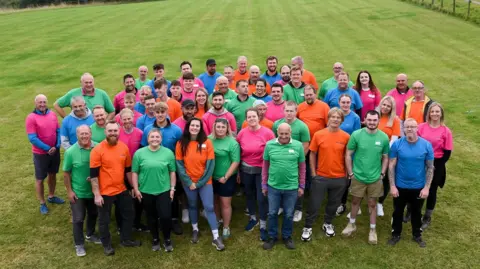 Pave-Aways A group of men and women in an assortment of brightly coloured plain green, orange, blue or pink t-shirts stand in a field looking up at a camera. They are all smiling.
