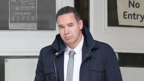 Eddie Mitchell A man with short black hair wearing a large dark blue coat. He is looking into and walking towards the camera.