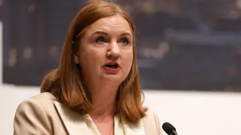Reuters A woman with red shoulder length hair talks into a microphone. She is wearing a cream and white jacket. 