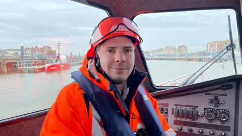 Ben Schofield/BBC Tom Duit wearing high visibility clothing, a life jacket and helmet, sat inside a Port of Lowestoft pilot vehicle