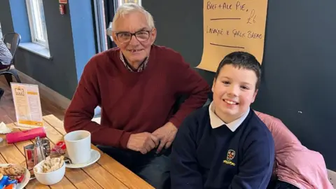 Dennis Spence, who is wearing a maroon jumper and patterned shirt, is sitting next to Jesse, who is wearing his school uniform. They are both smiling at the camera. 