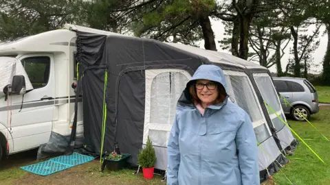 Penny Lidgard is wearing a blue jacket. She is standing in front of a white caravan.