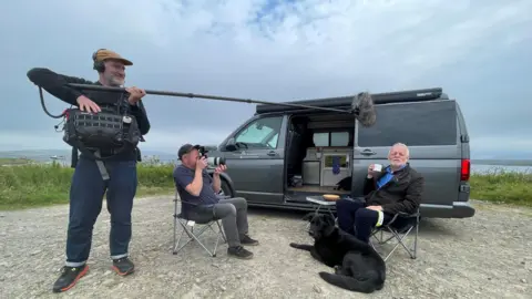Ian Hamilton and a BBC Scotland crew filming in Orkney. Ian and a cameraman are sitting in camping chairs in front of a grey camper van parked overlooking the Orkney coast. Another crew member, a man with a grey beard and a brown cap, is standing to the side, carrying recording equipment on a strap and holding a boom microphone. Major, the black cross Shepherd-Retriever guide dog, is lying on the ground in front of Ian.