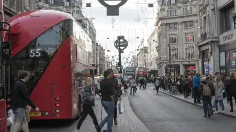 Getty Images Shoppers crossing Oxford Street at Christmas time including some people weaving between two buses, and a crowded pavement on the other side of the road