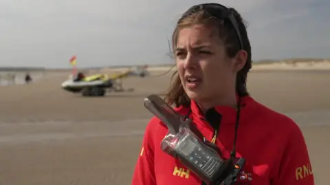 RNLI lifeguard Sarah Keast-Butler stands on Camber Sands beach wearing a red RNLI jacket and carrying a radio around her neck. Lifeboat equipment, sand and the shoreline can be seen behind her.