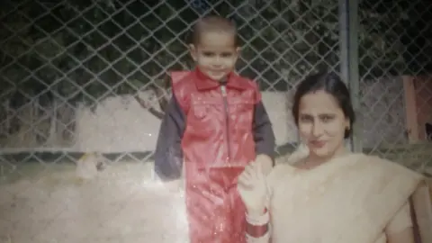 Tushar Kumar A slightly faded photograph shows a small child (Tushar Kumar) in a bright red outfit standing beside an woman near a chain-link fence. The adult wears a light-coloured outfit and jewellery, while the child’s shiny red clothing stands out against the muted background. Behind them, a light-coloured animal can be seen inside the enclosure. 