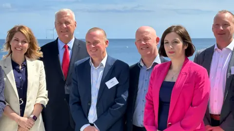 Local politicians including, North East Mayor Kim McGuiness (first left) are all dressed in suit jackets of different colours including cream, beige, blue and pink. They are standing close together with their hands crossed in front of the North Sea at the Energy Central Learning Hub in Blyth.