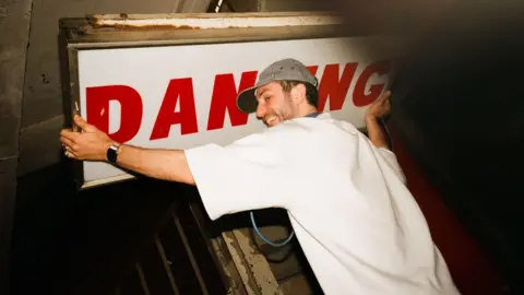 A man in a white t-shirt and stripy hat is hugging a white sign reading 'Dancing' in red writing. He appears to be in a warehouse.