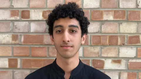 A young man with a black shirt looks at the camera and stands against a brick wall 