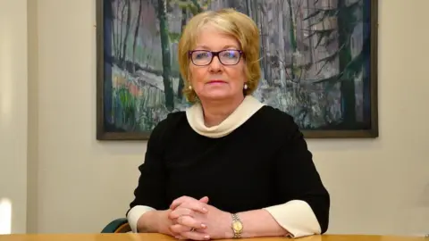 Pacemaker Marie Anderson is a middle-aged woman with blond hair and black-rimmed glasses. She is wearing a black jumper with white sleeves and neck and is at a desk with a a painting of trees in the background