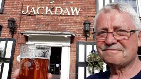 A man with short white hair and a white moustache is wearing glasses while holding up a pint of beer. A red brick pub behind him is called Jackdaw. 