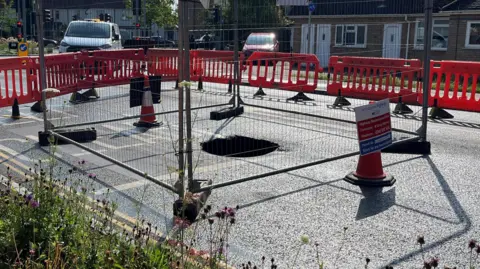 A sinkhole is in the middle of a road which has been closed. Around the sinkhole is a metal fence and cones with signs. Surrounding that are red barriers which have closed the road.
