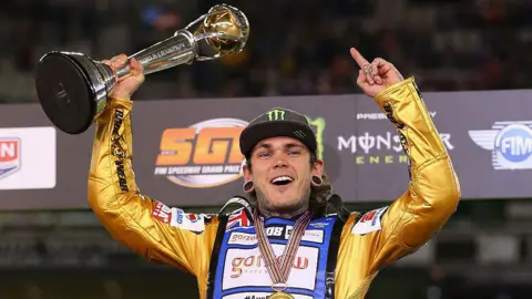 Quinn Rooney/Getty Images A speedway rider wearing a gold racing suit holds a trophy in the air. A display featuring championship branding and sponsor logos is behind him.