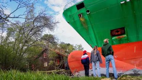 Three people, wearing jeans and casual jackets, stand and stare up at a huge green container ship which has run aground on land. A house looks small and is next to the ship.