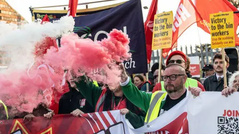 Getty Images A large protest. A man can be seen holding a banner. He has a bald head and is wearing glasses. He is holding a red flare. There are people all around him holding placards. They look suitably determined.