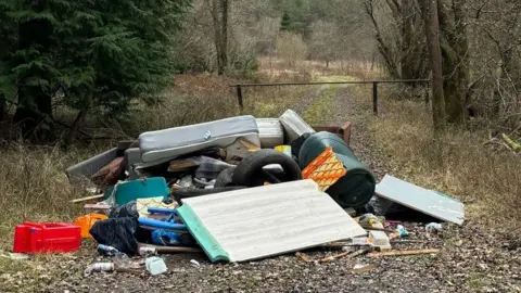 A large pile of discarded waste dumped along an overgrown pathway in Bentley Woods. There is a large section of lino flooring visible, a tyre, multiple mattresses, a green plastic barrel and other household items.