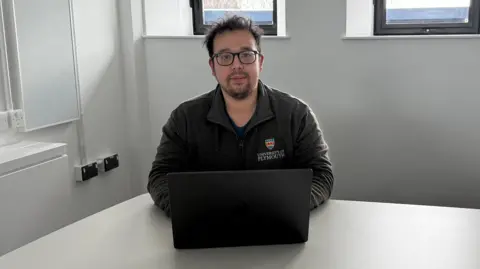 Lecturer Dr Ben Siu sitting at white table in front of a laptop, looking at the camera wearing a University of Plymouth fleece