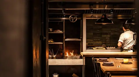 Eleonora_Boscarelli Chef Corrin with seen from behind in white uniform preparing food in open kitchen. Tables laid for guests are visible just on the other side and to his left are shelves and a small open fire for cooking.