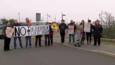 Protestors stand outside the incinerator in a line in Barry Docks. They are holding signs in protest, with the most visible being a sign saying 'No incinerator, no toxic ash'.