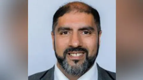 Worcester City Council A man smiles against a grey background. He has dark hair and greying beard. He is wearing a dark jacket and white shirt.