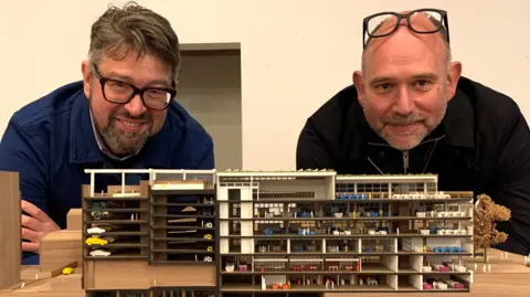 Two men wearing dark coloured jumpers and glasses lean on a desk behind a scale model of the former John Lewis store