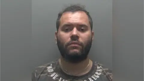 Cambridgeshire Constabulary Custody photo of Daniel Barbosa against a grey background. He has short dark hair and facial hair. He is wearing a gold chain and camouflage style top with writing around the neck.