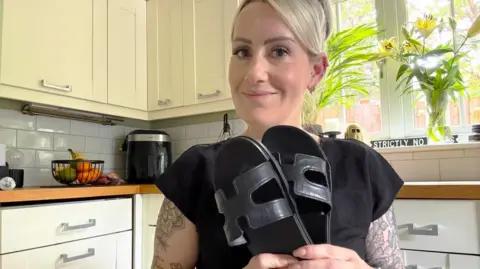 Emma Neil with blonde hair in a kitchen holding her sandals