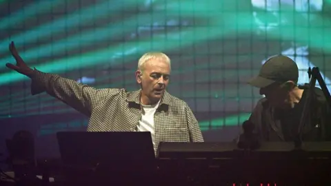 Karl Hyde of Underworld performing. He has short, blond hair and is wearing a checked jacket. There are green lights illuminating the back of the stage