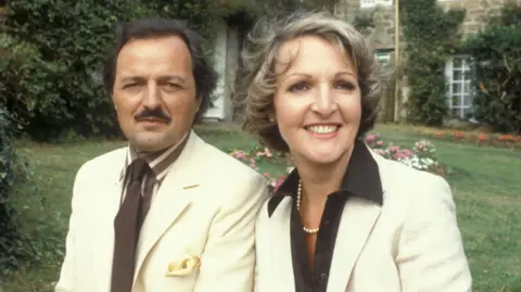 BBC Archive image of a man and a woman sat next to each other in front of a stately home. They are wearing posh clothes in a suit and tie and formal attire.
